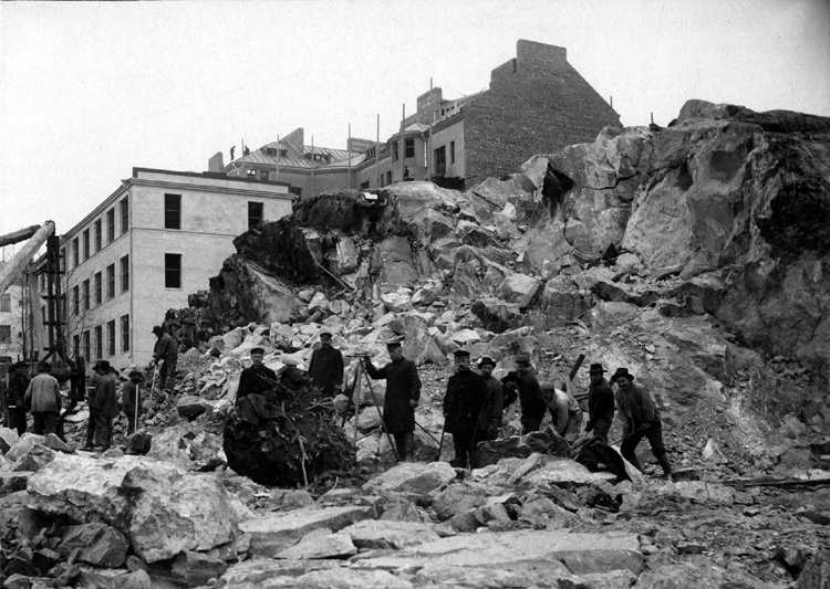 Black and white photograph of a surveyor and workmen beside the granite outcrop.